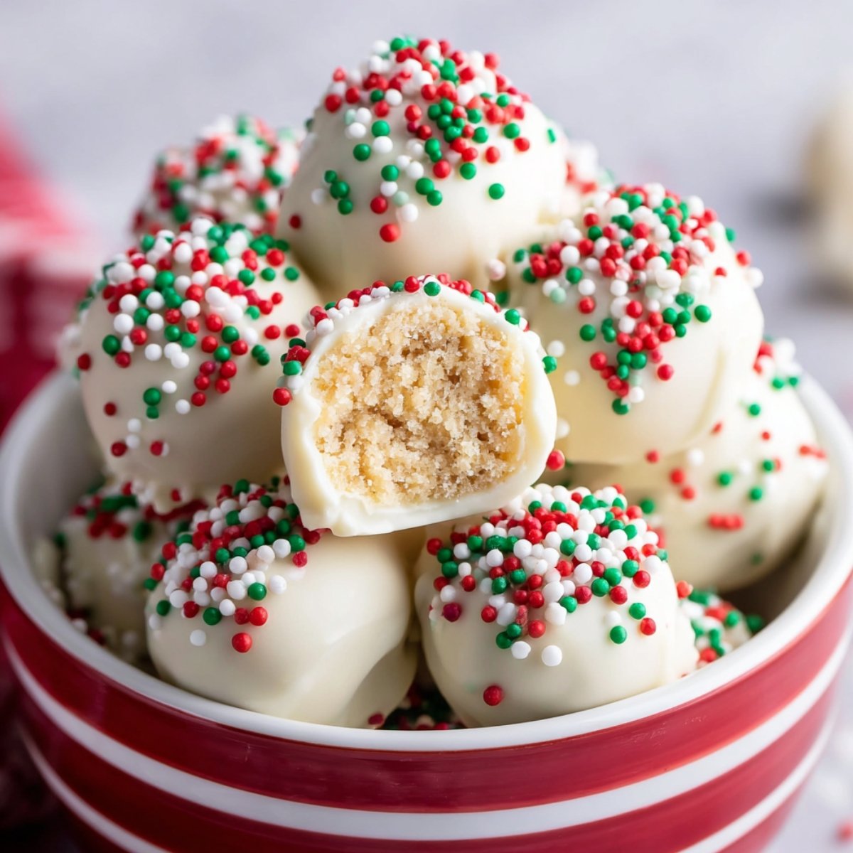 Sugar cookie truffles covered in white chocolate and colorful holiday sprinkles, with one truffle broken open to show the crumbly filling inside.