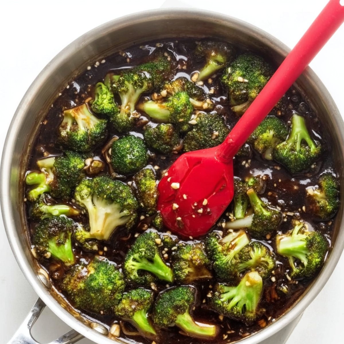 Broccoli florets simmering in a thick, dark honey garlic sauce in a skillet, with visible garlic pieces and a red spatula stirring the mixture.