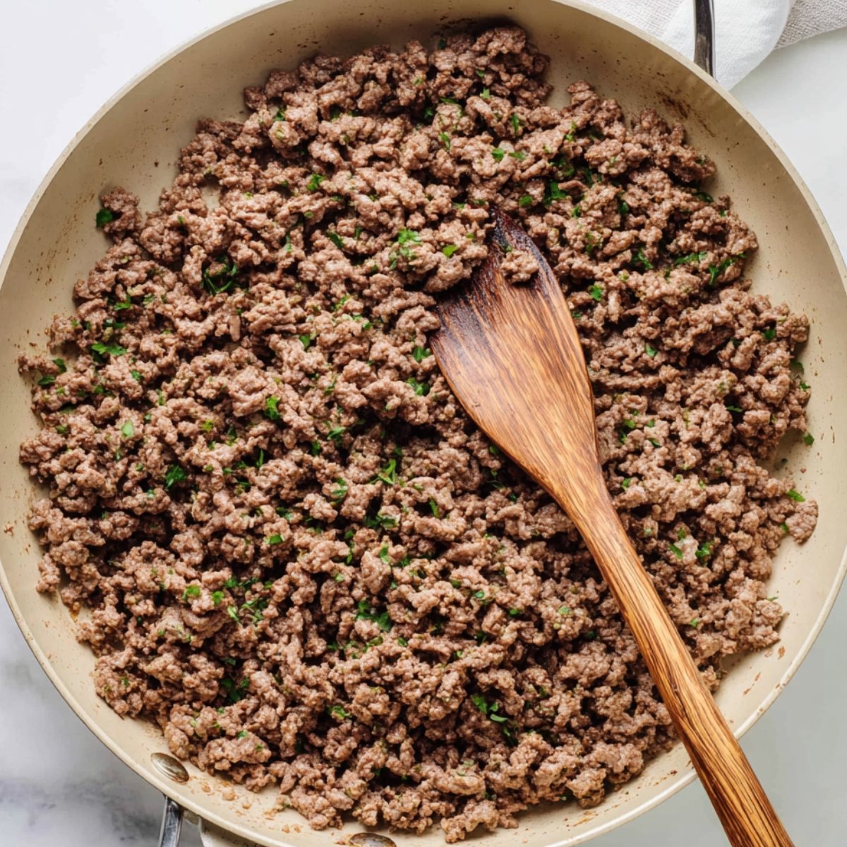 Overhead view of browned ground pork cooking in a skillet with a wooden spoon and chopped herbs