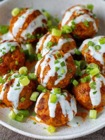A close-up shot of cooked Buffalo Chicken Meatballs covered in spicy buffalo sauce, garnished with chopped green onions and served in a bowl.