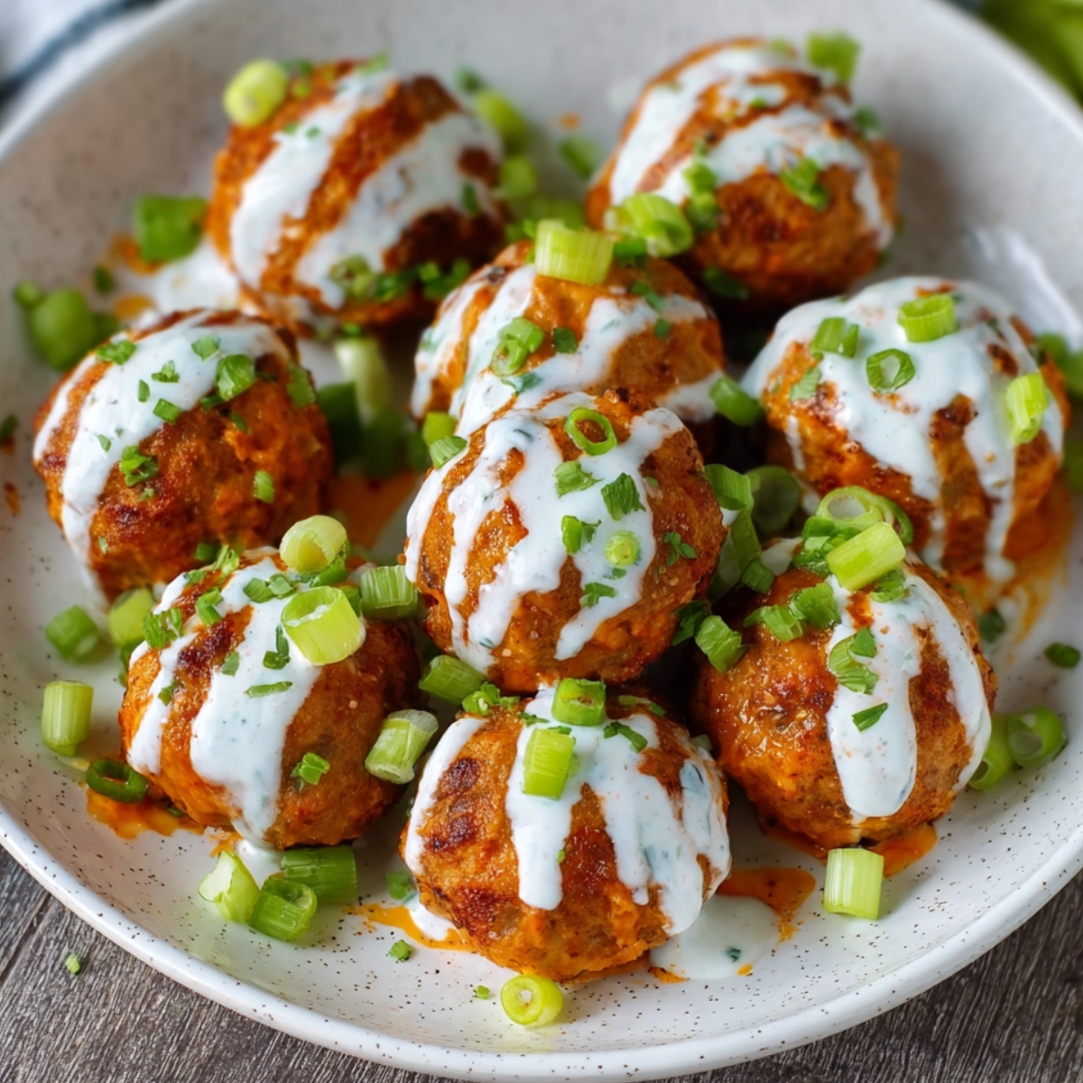 A close-up shot of cooked Buffalo Chicken Meatballs covered in spicy buffalo sauce, garnished with chopped green onions and served in a bowl.
