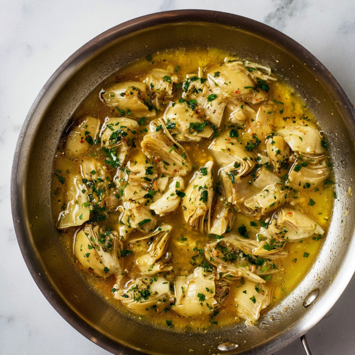 Artichoke hearts simmering in a lemon butter garlic sauce with parsley in a stainless steel skillet, top-down view on marble background.