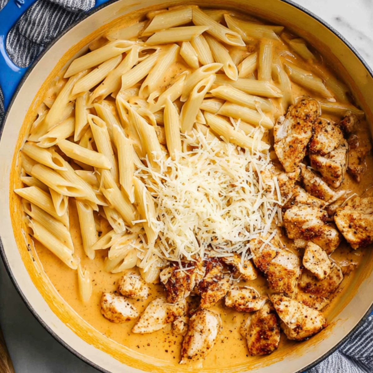 Penne pasta, Cajun-seasoned chicken, and grated parmesan added to creamy sauce in a skillet before mixing.