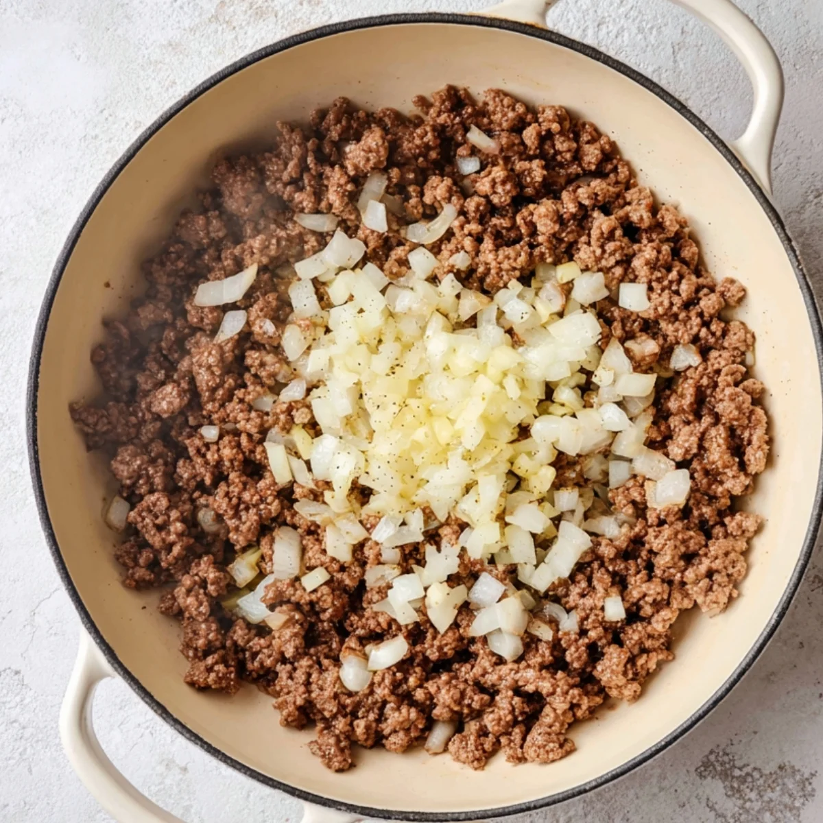 Browned ground beef in a skillet topped with diced onion and garlic before mixing, overhead cooking step