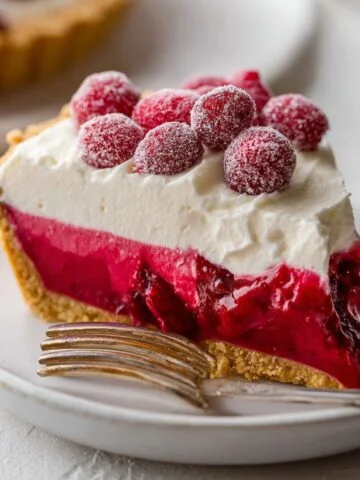 Slice of Cranberry Curd Tart topped with whipped cream and sugared cranberries on a plate with a fork.