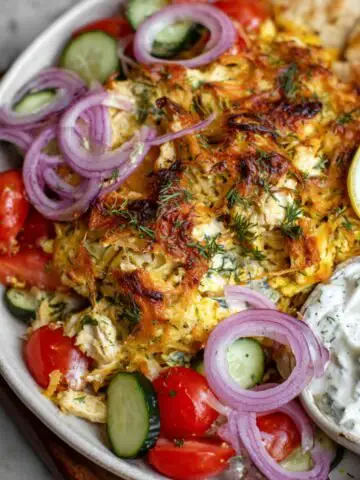 A close-up view of a Chicken Tzatziki Casserole served with a side of rice, garnished with onions, lemon wedges, and a creamy white sauce.