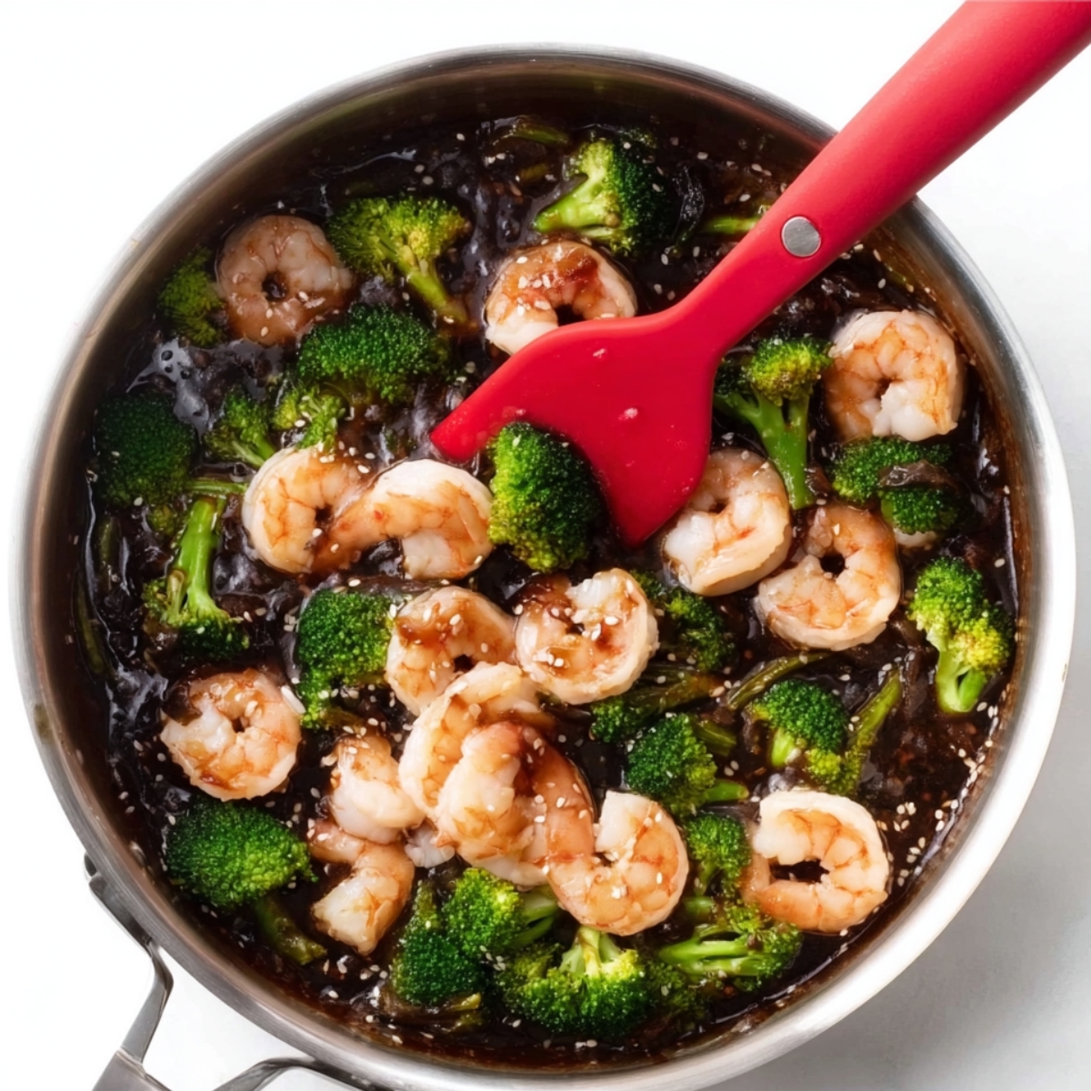 Raw shrimp being added to a skillet filled with broccoli and glossy honey garlic sauce, stirred with a red silicone spatula during the cooking process.