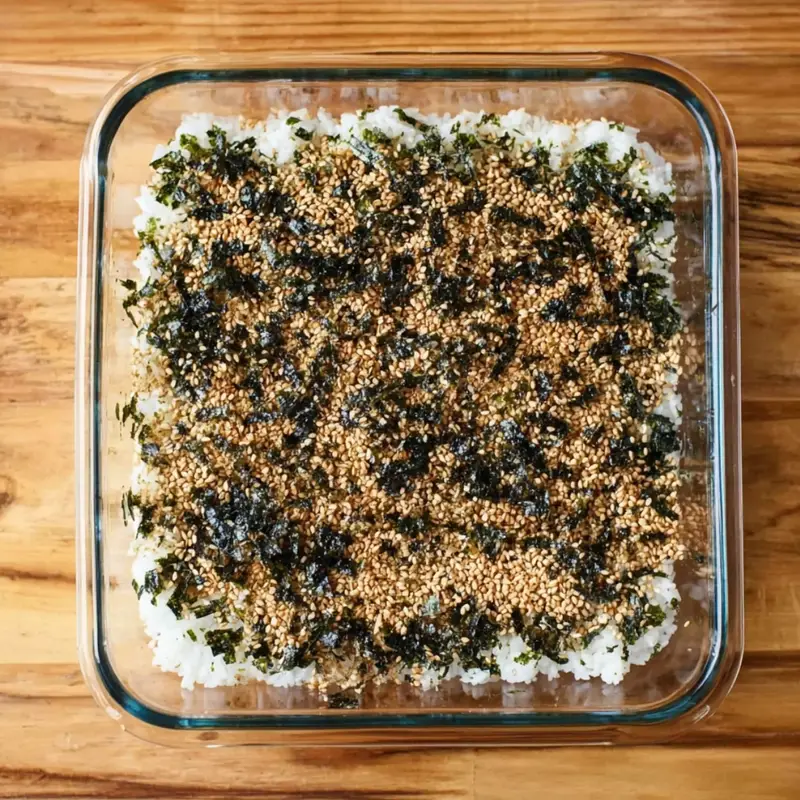 Press the seasoned sushi rice evenly into the bottom of the prepared baking pan. Use the back of a spoon or your rice paddle to pack it down gently. Sprinkle the furikake seasoning over the rice for extra flavor and crunch.