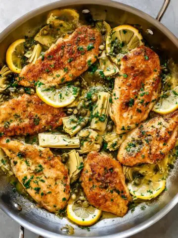 Lemon artichoke chicken in a skillet with golden seared chicken breasts, artichoke hearts, capers, parsley, and lemon slices in a buttery sauce.