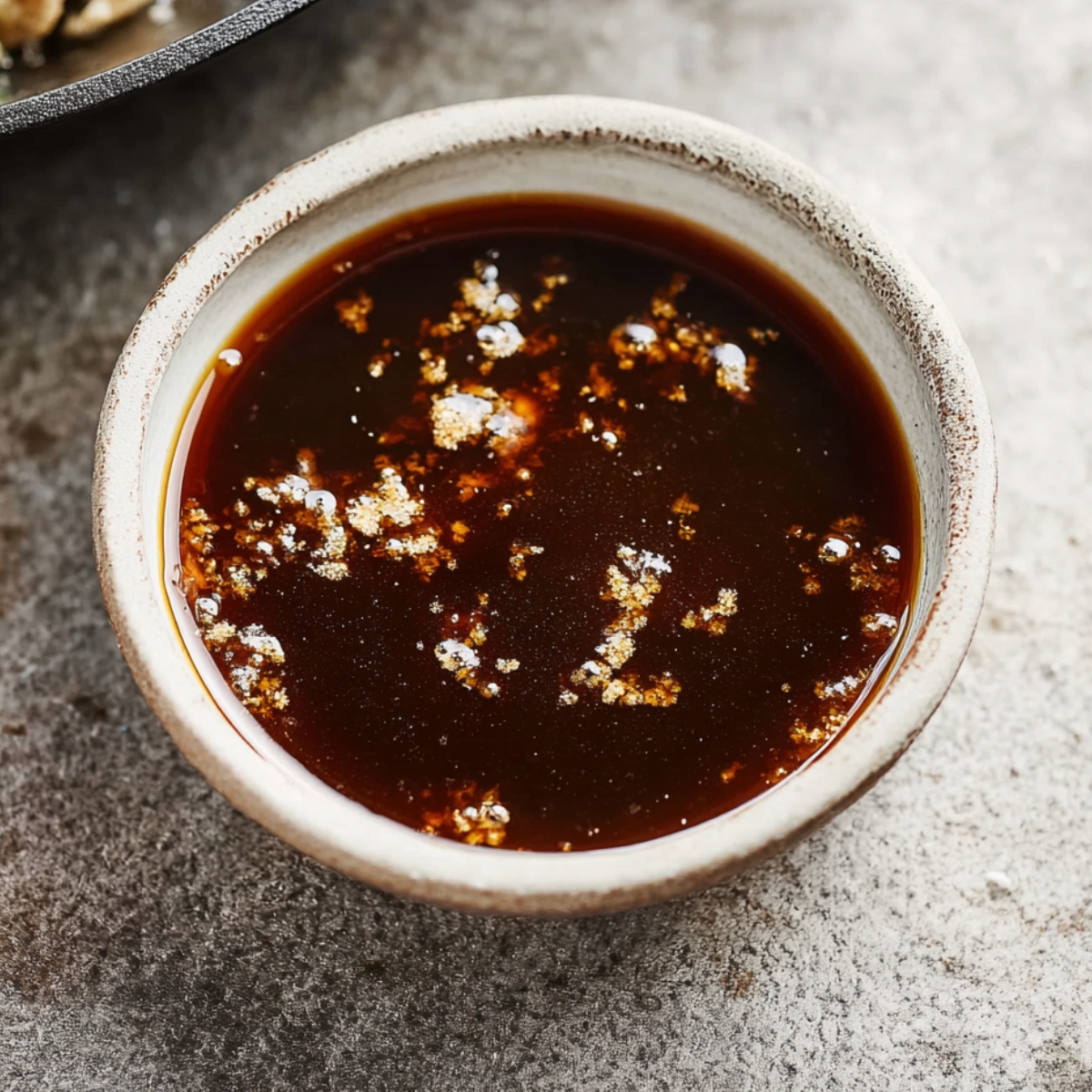 A small bowl filled with a dark, glossy sauce, likely made with soy sauce or a similar base, with small bits of seasoning visible floating on the surface.