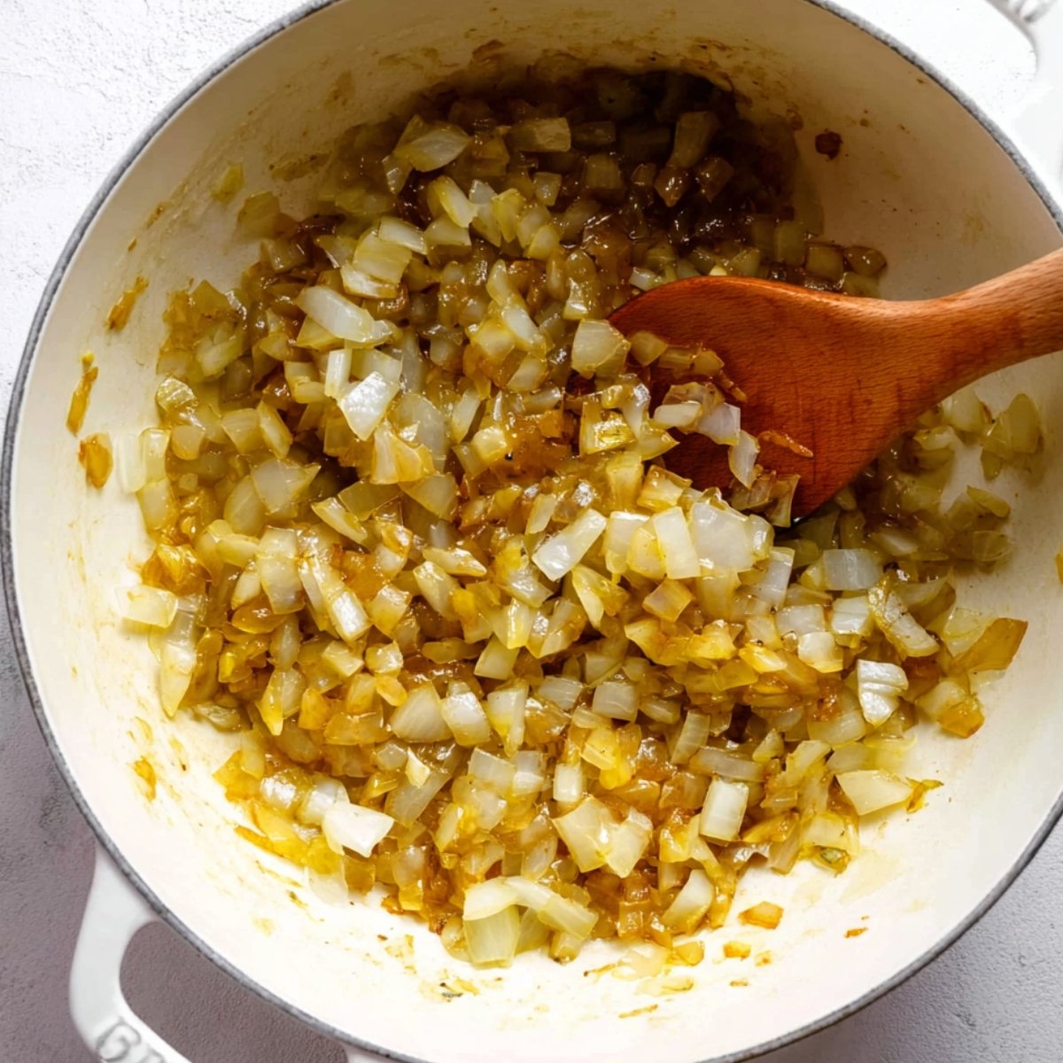 Golden sautéed diced onions cooking in a white pot with a wooden spoon.