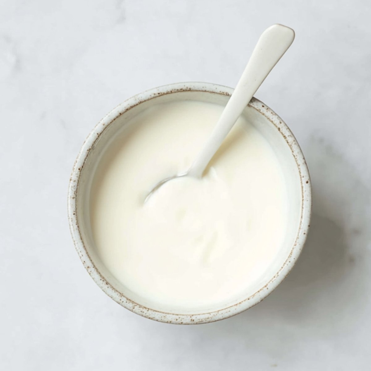 Small ceramic bowl filled with smooth vegan sour cream and a spoon, photographed from above on a light marble surface.