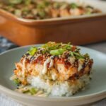 A close-up of Spicy Salmon Sushi Bake on a plate, topped with sesame seeds, green onions, and creamy sauce, with a baking dish in the background.