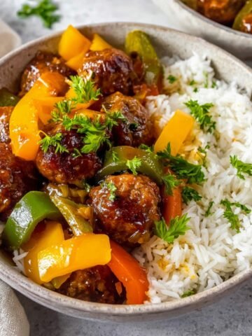 Sweet and sour meatballs served with colorful peppers, garnished with fresh parsley on a bed of rice.