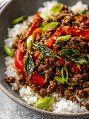 A completed dish of Thai basil beef served over white rice in a gray bowl. The dish features ground beef, red bell peppers, and fresh basil leaves, garnished with sliced green onions.