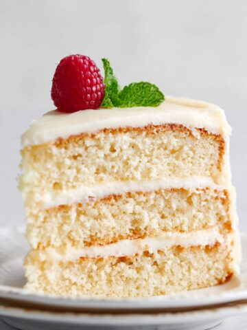 A slice of Vanilla Cake showing the layers of soft cake and frosting, topped with a raspberry and mint sprig.