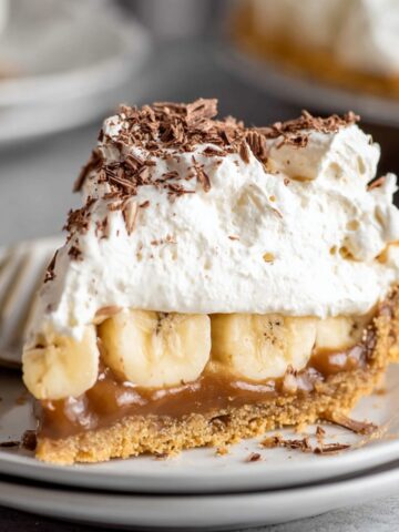 A close-up of a decadent slice of Banoffee Pie, topped with whipped cream, chocolate shavings, and a layer of banana slices on a buttery graham cracker crust.