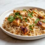 A plate of delicious Chicken Pulao, garnished with fresh cilantro, served with a side of cucumber slices and yogurt.