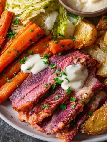A plate of sliced Corned Beef and Cabbage served with roasted potatoes, glazed carrots, and sautéed cabbage, topped with a creamy sauce and garnished with parsley.
