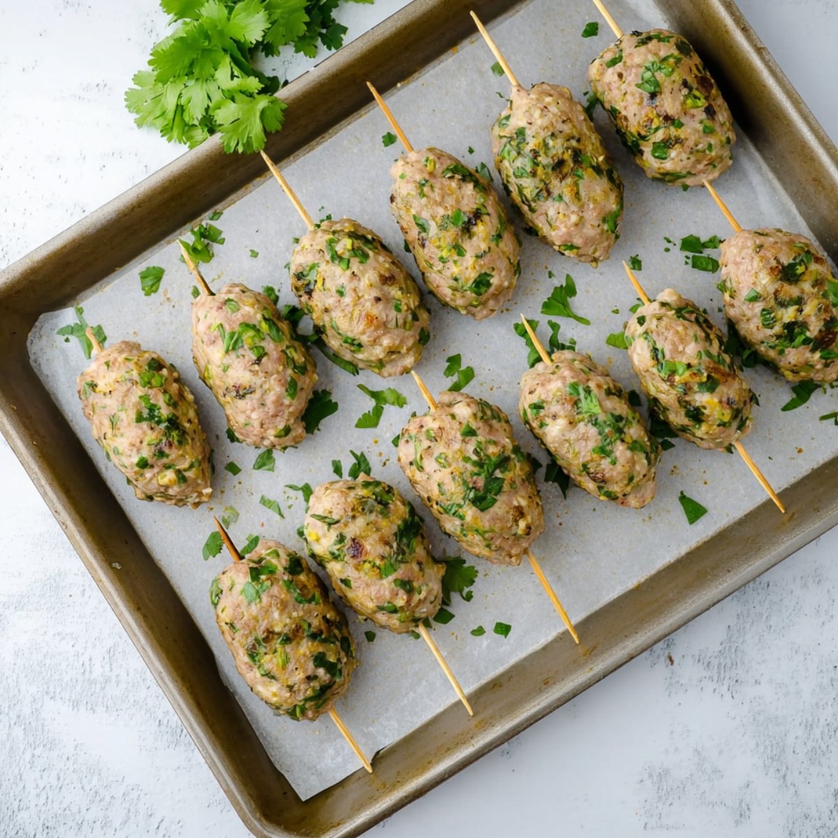 Freshly formed chicken kofta kebabs placed on a parchment-lined baking tray, ready to be grilled or baked, with chopped cilantro adding a vibrant green color.