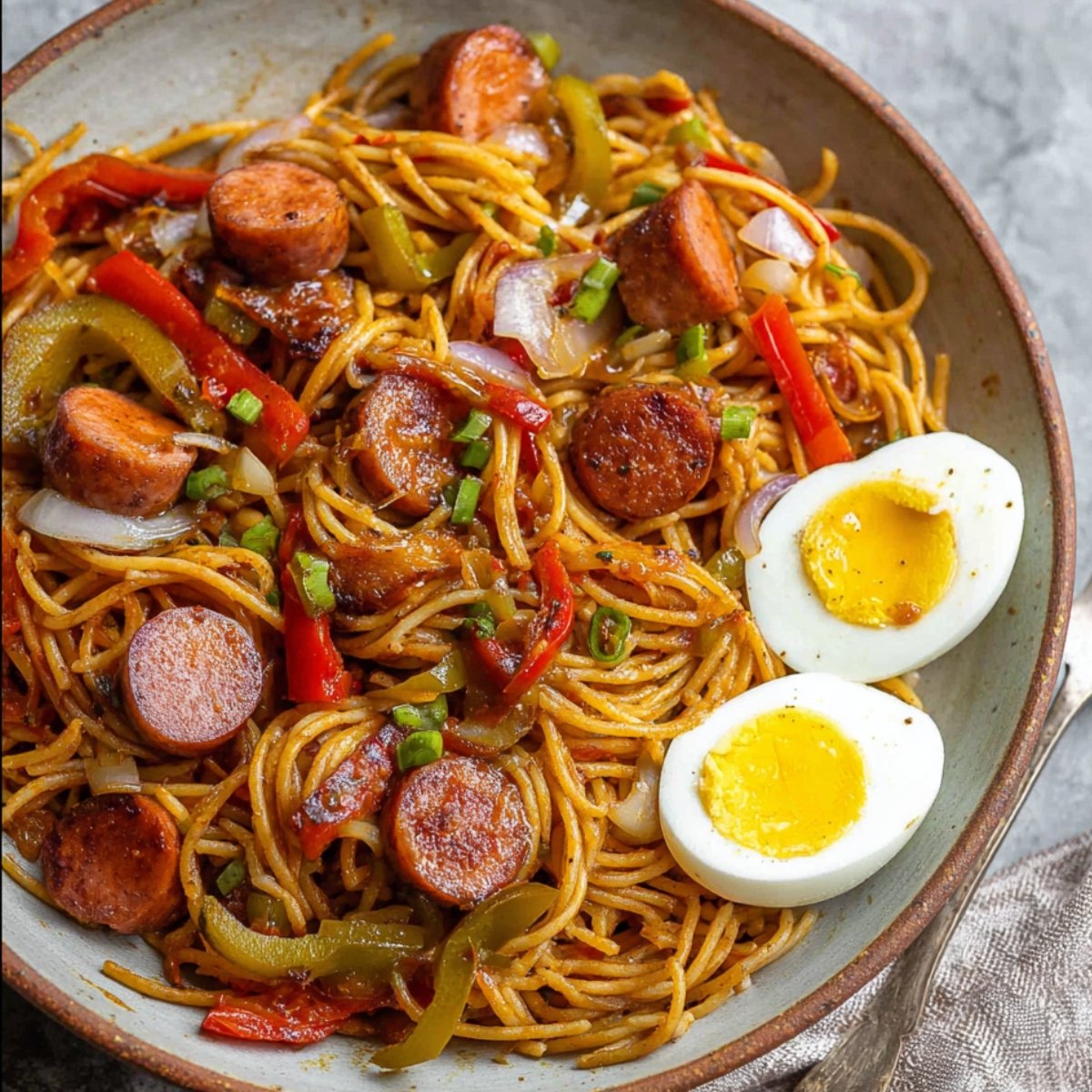 A serving of Haitian spaghetti in a rustic bowl with sliced sausages, bell peppers, onions, and soft-boiled eggs on the side. The dish is colorful and hearty, making for a comforting meal.