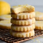 A stack of Lemon Shortbread Cookies with a glaze and lemon zest topping, one cookie with a bite taken out, freshly baked and cooling on a wire rack.