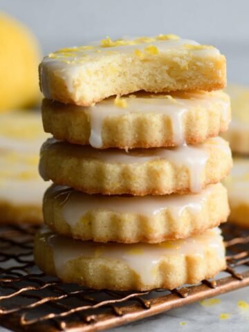 A stack of Lemon Shortbread Cookies with a glaze and lemon zest topping, one cookie with a bite taken out, freshly baked and cooling on a wire rack.