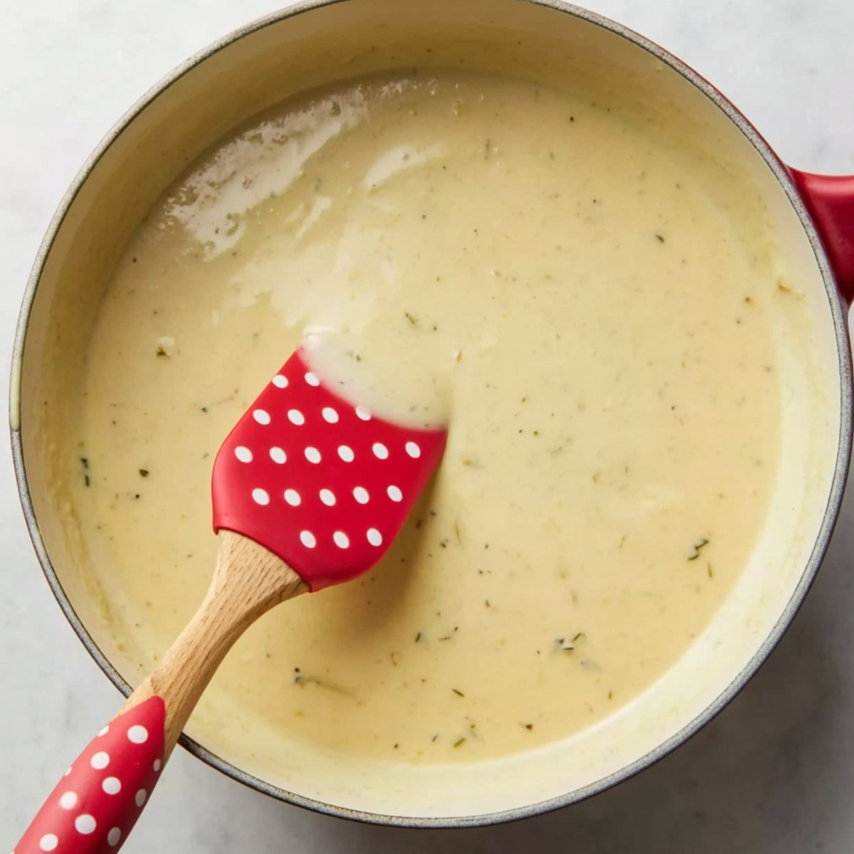 A creamy, smooth ranch sauce in a pan, being stirred with a red spatula with white polka dots. The sauce has a rich, velvety texture and visible herbs mixed in.