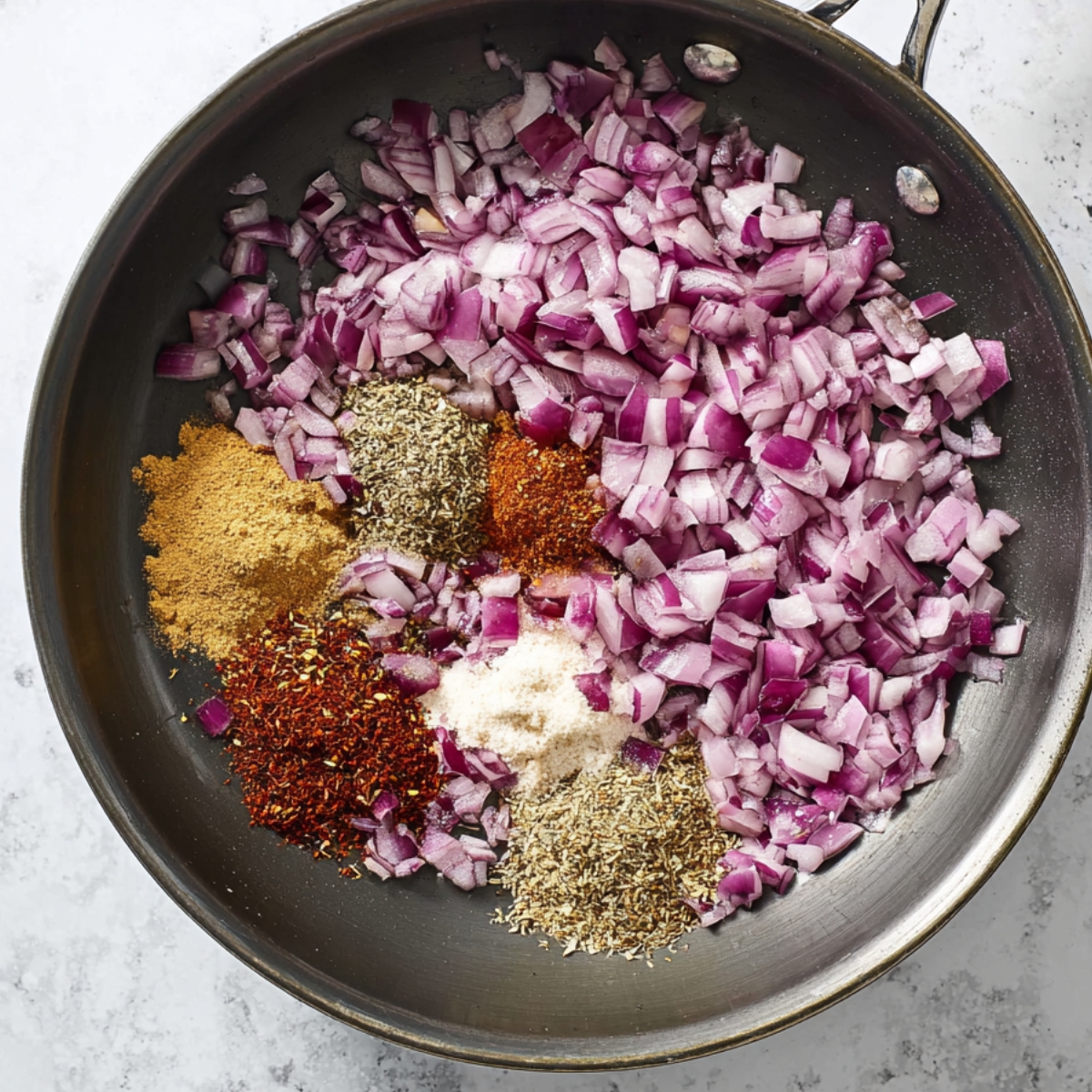 A skillet with finely chopped red onions and a variety of spices like cumin, chili powder, garlic powder, and oregano, all ready to be sautéed together for added flavor.
