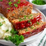 A plate of Vegan Lentil Loaf slices, with mashed potatoes and green peas, topped with parsley and glazed with ketchup.