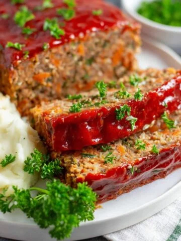 A plate of Vegan Lentil Loaf slices, with mashed potatoes and green peas, topped with parsley and glazed with ketchup.