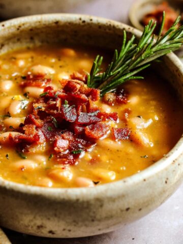 A warm bowl of White Bean and Bacon Soup, topped with crispy bacon bits and a sprig of fresh rosemary for extra flavor and garnish.