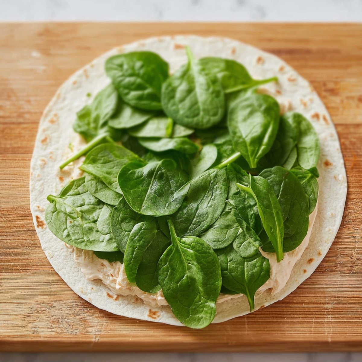 A close-up of a tortilla spread with creamy filling and topped with fresh spinach leaves on a wooden cutting board.