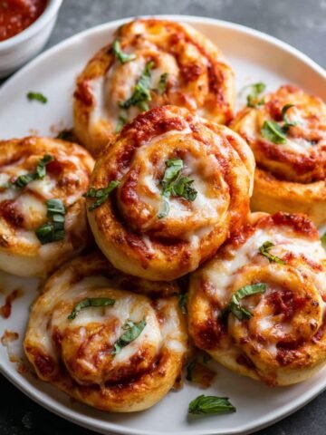 A plate of golden, baked Homemade Pepperoni Pizza Rolls, with melted cheese, marinara sauce, and fresh basil, perfect for serving as a snack or appetizer.