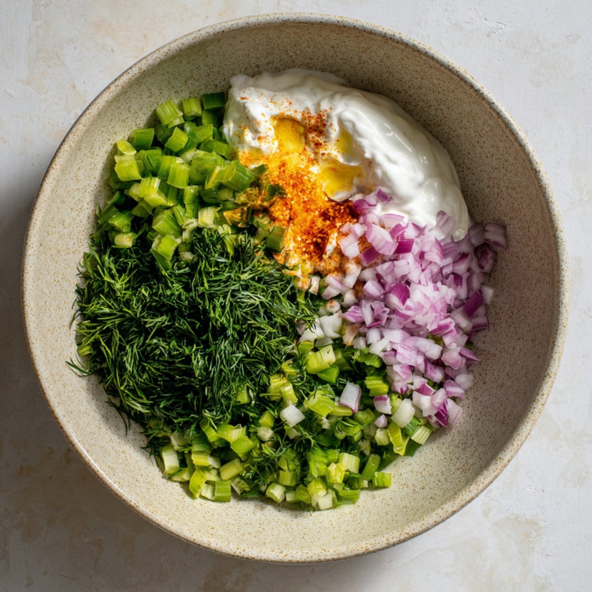 Ingredients for a sauce including yogurt, diced celery, red onions, and spices, mixed together in a bowl.