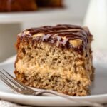 A slice of peanut butter banana cake with a layer of peanut butter filling, topped with a glossy layer of chocolate ganache, showcasing the moist texture of the cake and the rich filling.