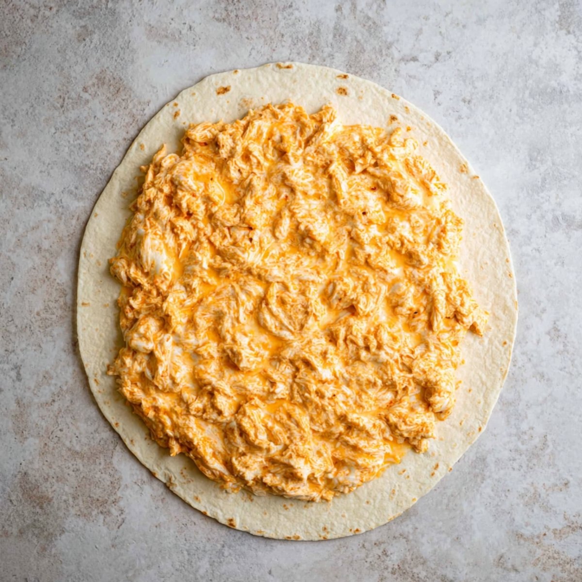 A tortilla with buffalo chicken mixture spread on it, ready for rolling.